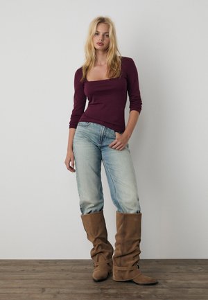 Donna con capelli biondi lunghi che indossa un top color vino con scollo quadrato, jeans azzurri e stivali marroni arricciati al ginocchio, in piedi su pavimento di legno.