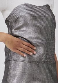 Metallisk sølvfarvet strapless top med et tekstureret, diagonalt mønster. Nærbillede viser en hånd, der hviler på stoffet, hvilket fremhæver dets glans og struktur.