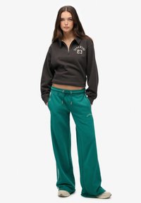 Felpa nera a maniche lunghe, cropped, con collo zip e logo, abbinata a pantaloni larghi teal con coulisse e dettagli di branding.