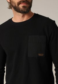 Chemise noire à manches longues avec une texture côtelée et une poche carrée sur le côté gauche de la poitrine. L'étiquette sur la poche indique "DEELUXE."