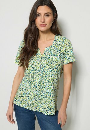 Kobieta o długich ciemnych włosach, ubrana w krótki rękaw, kwiecistą bluzkę z plisami i guzikami, w połączeniu z niebieskimi dżinsami, stojąca na tle białej ściany.