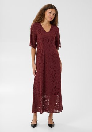 Femme aux cheveux bouclés portant une robe midi en dentelle bordeaux avec des manches aux coudes et des talons noirs pointus, debout devant un fond uni.