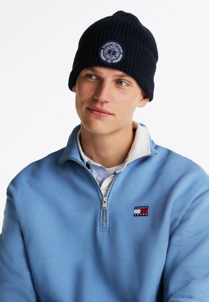 Marineblauwe gebreide beanie met een voorste logo patch; gedragen met een lichtblauwe rits-sweater met een logo op de borst en een overhemd eronder.