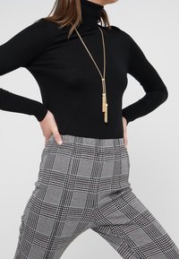 Maglione nero a collo alto realizzato in tessuto morbido, abbinato a pantaloni a vita alta in tessuto a scacchi grigi e bianchi con dettagli neri. Dettaglio della collana in oro.