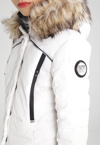 Bílá péřová bunda s kapucí z umělé kožešiny, se zipovými kapsami, černými akcenty a kulatou nášivkou na rukávu. Strukturovaná prošívaná tkanina.