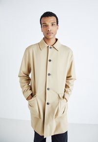 Beige esőkabát, gallérral és öt elülső gombbal, oldalsó zsebekkel és laza fazonnal. Simaság és minimalista dizájn jellemzi.