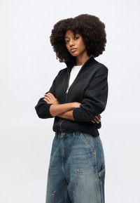 Μαύρο πουλόβερ με φερμουάρ, με ριμπ μανσέτες και γιακά, συνδυασμένο με ανοιχτό μπλε τζιν παντελόνι με φαρδιά πόδια, μεγάλα τσέπες και ξεβαμμένη όψη.