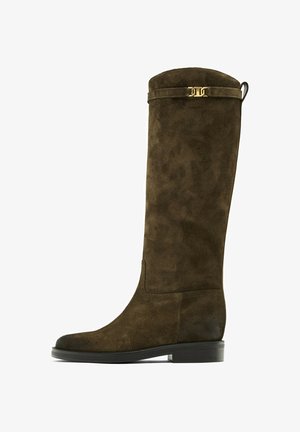 Bottes hautes en suède brun avec un bout arrondi, une semelle plate et un accent en métal doré fin sur le dessus, présentant une texture douce et un design élégant.
