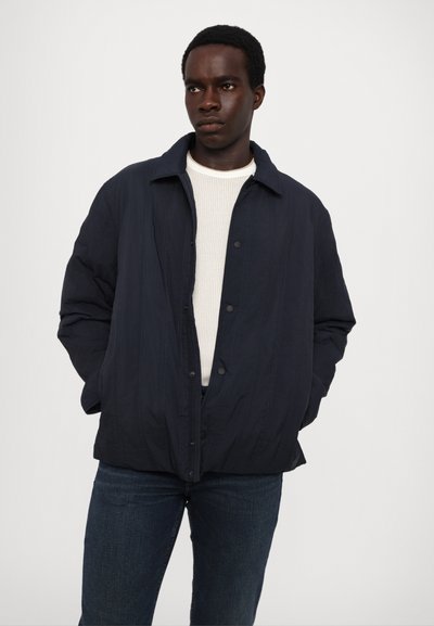 Marineblaue gesteppte Jacke mit Knopfleiste, spitzem Kragen und Seitentaschen, kombiniert mit einem weißen gerippten Pullover und dunklen Jeans.