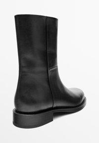Bottine en cuir noir avec une texture lisse, un bout arrondi et une semelle en caoutchouc plate. Présente un minimum de couture et une silhouette épurée.