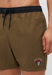 Khaki plavkové šortky s čiernym elastickým pásom, ktorý má šnúrkový uzáver a farebnú vyšívanú nášivku s lebkou "Santa Cruz".