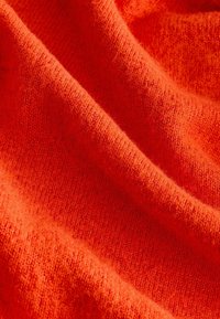 Gros plan d’un tissu tricoté doux de couleur orange vif avec une texture visible et des plis doux créant ombres et reflets.