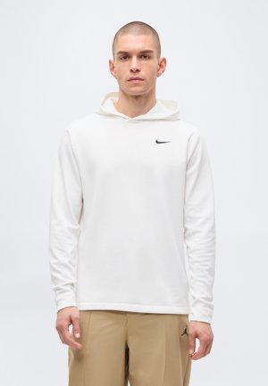 Man met kort haar, gekleed in een witte Nike-hoodie en beige broek met een Jordan-logo, staand tegen een effen achtergrond.