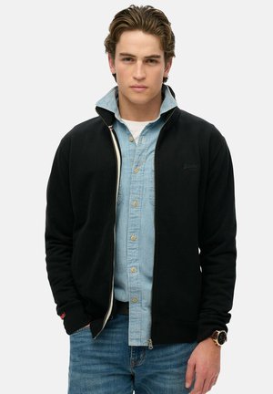 Felpa nera con zip e colletto alto, indossata sopra una camicia azzurra a bottoni. Presenta polsini a costine e un logo sul petto.