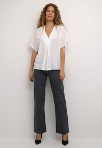 Donna con capelli ricci che indossa una blusa bianca a maniche corte, jeans scuri grigi decorati e tacchi neri a punta, su uno sfondo semplice.