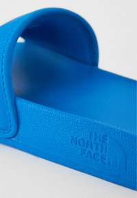 Modra gumijasta drsna obutev z teksturirano površino, zaobljenim paščkom in vtisnjenim logotipom "The North Face" na podplatu.
