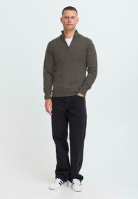 Man die staat met de hand in zijn zak, gekleed in een olijfgroen trui met rits, zwarte broek en witte sneakers met zwarte strepen op een witte achtergrond.
