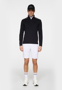 Svart långärmad tröja med fjärdedels dragkedja, vita detaljer, kombinerad med vita shorts. Tillbehören inkluderar en svart keps och vita strumpor med svarta ränder.