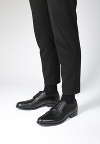Zwarte leren dresschoenen met een wingtip-design, brogue-details en veters, gecombineerd met zwarte broeken en sokken.