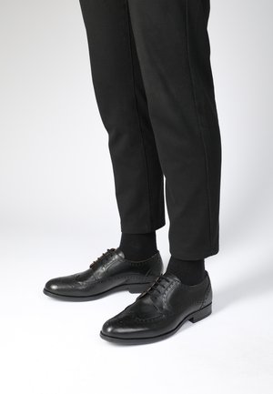Černé kožené společenské boty s wingtip designem, brogue detaily a šněrováním, v kombinaci s černými kalhotami a ponožkami.