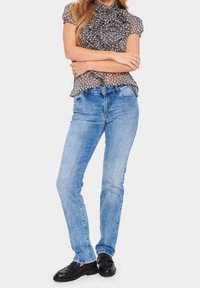 Donna che indossa jeans dritti blu chiaro, mocassini neri e una blusa a maniche corte con volant e stampa leopardata, in piedi con le braccia incrociate.