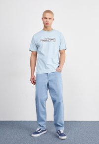 Camiseta de algodón azul claro con el logo negro "VOLCOM", combinada con pantalones de mezclilla azul claro y zapatillas azules sobre un suelo texturizado.