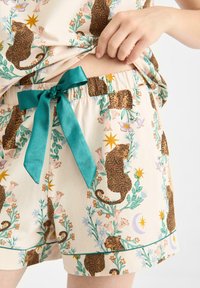 Krátké pyžamové šortky v světle béžovém materiálu, s leopardím a květinovým vzorem, s tyrkysovou saténovou mašlí v pase a zeleným lemem.