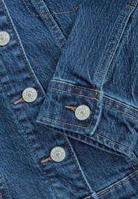 Denim fabric in dark blue with a smooth texture. Features silver metal buttons with embossed text, contrasting orange stitching, and a layered cuff.