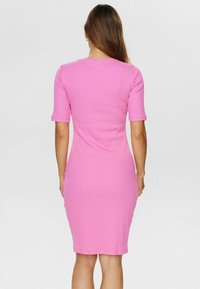 Femme avec de longs cheveux portant une robe rose ajustée, arrivant au genou, à manches courtes, se tenant de dos devant un fond clair.