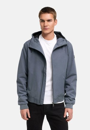 Jeune homme portant une veste à capuche bleue zippée sur un t-shirt blanc et un pantalon noir, debout devant un fond clair uni.