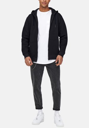 Schwarze Zip-Hoodie mit einer Kordelzugkapuze und Fronttaschen, kombiniert mit einem weißen T-Shirt und grauen Denim-Jeans. Weiße Sneakers vervollständigen das Outfit.