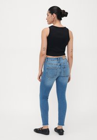 Black crop tank top with blue skinny jeans. Jeans feature a frayed hem, back pockets, and a tan leather logo patch at the waistband.