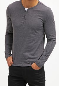 Mężczyzna ubrany w dopasowaną, ciemnoszarą bluzę henley z długim rękawem i czterema guzikami oraz czarne spodnie, z rękami w kieszeniach.