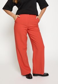 Rote weite Hosen aus glattem Stoff, mit hoher Taille, zwei seitlichen Taschen und geradem Schnitt. Stylisch kombiniert mit einem schwarzen Oberteil.