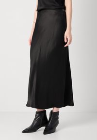 Svart satin midi-nederdel med en glat tekstur, løs pasform og subtil slids i siden, parret med sorte spidse ankelstøvler.