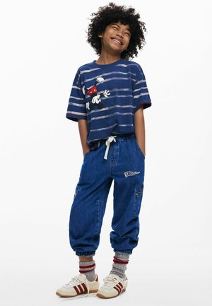 Nasmejano dijete s kovrčavom kosom koje nosi prugastu tamnoplavu majicu s motivom Mickey Mousea, široke traperice, sive čarape i bijele tenisice s crvenim prugama.