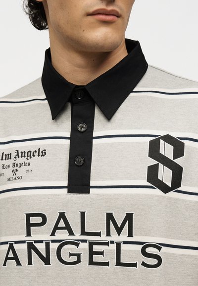 Άνδρας φοράει γκρι και λευκή ριγέ πόλο μπλούζα με μαύρο γιακά, πατιλέτα με κουμπιά και έντονο κείμενο "Palm Angels" καθώς και μαύρο λογότυπο στο στήθος.