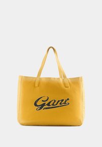 Kollane kott, millel on suur must retrostiilis "Gant" logo, ristküliku kuju, kangasmaterjal ja kaks lühikest rihma.