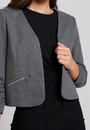 Femme portant un blazer court gris texturé avec des poches zippées sur une robe noire côtelée à devant torsadé, main tenant le revers du blazer.