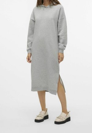 Kvinde iført en lang grå hættetrøjekjole med sideslids og cremefarvede frynset loafers på hvid baggrund.