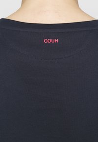 Marinblå t-shirt med rund halsringning. Har en röd "HUGO"-logotyp på baksidan. Tillverkad av mjukt, lätt tyg.