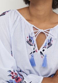 Světle modrá pruhovaná blůza s květinovou výšivkou v červené a modré, s uvazováním na krku a třásněmi. Dlouhé rukávy s nařasenými manžetami.