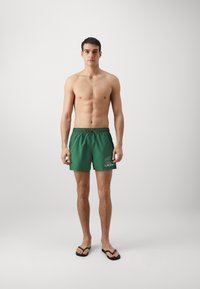 Grønne svømmedrakter laget av lett stoff, med elastisk midjebånd, snor og "LACOSTE"-logo i hvitt. Svarte flip-flops.