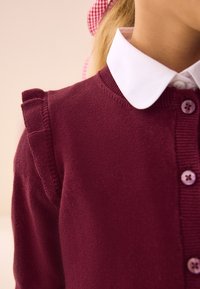 Bordeauxrode gebreide cardigan met geruite schouderaccenten en daaronder een wit overhemd met kraag. De knopen hebben dezelfde kleur als de cardigan.