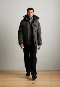 Giacca isolata verde scuro con molteplici tasche, dettagli con zip e cappuccio regolabile, indossata sopra una maglietta grigia e pantaloni neri, in piedi su un pavimento in legno.