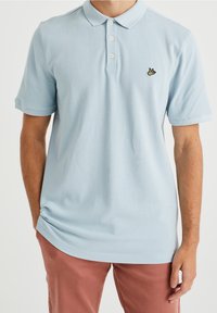 Polo shirt bleu clair avec un tissu texturé, patte de boutonnage à deux boutons et un petit logo brodé noir et jaune sur la poitrine.