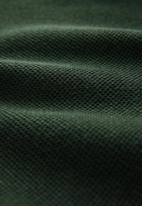 Textured dark green fabric with a mesh-like pattern, featuring soft, uneven folds that create depth and subtle shading.