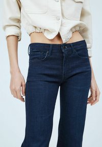 Tummansiniset denimpohjat, joissa on muotoiltu vyötärö, klassinen nappikiinnitys ja huomaamattomat ompeluyksityiskohdat; yhdistettynä vaaleanbeigeen paitaan.