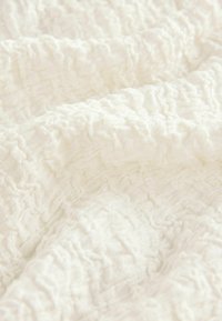 Weißer, strukturierter Stoff mit einem zerknitterten Muster, das weiche, gewebte Fasern zeigt und Tiefe sowie Dimensionen über die Oberfläche schafft.
