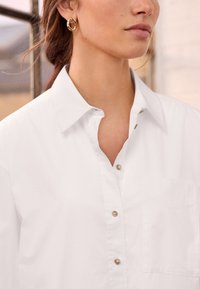 Hvid skjorte med knapper og spidse krave, frontal lomme og subtil glans, parret med guldrunde øreringe.